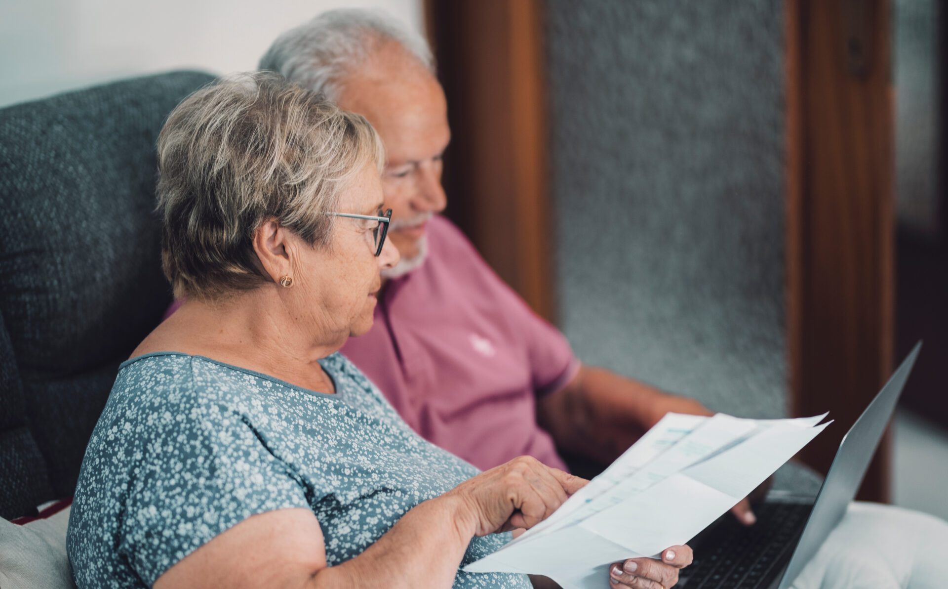 SIde view of real senior couple at home paying bills online with old-couple-sitting-sofa-during-video-call-tablet-old-couple-waving-tablet-planning-home